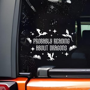 May include: White vinyl decal with the text "PROBABLY READING ABOUT DRAGONS" on a black background. The design includes dragon silhouettes, stars, and open books. The decal is applied to a vehicle window.