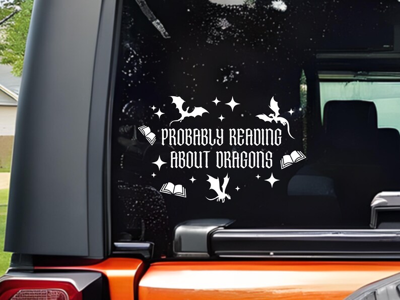 May include: White vinyl decal with the text "PROBABLY READING ABOUT DRAGONS" on a black background. The design includes dragon silhouettes, stars, and open books. The decal is applied to a vehicle window.