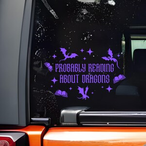 May include: Purple vinyl decal on a vehicle window, featuring the text "Probably Reading About Dragons" in a decorative font. The design includes dragon and book illustrations. The decal is applied to a black window.