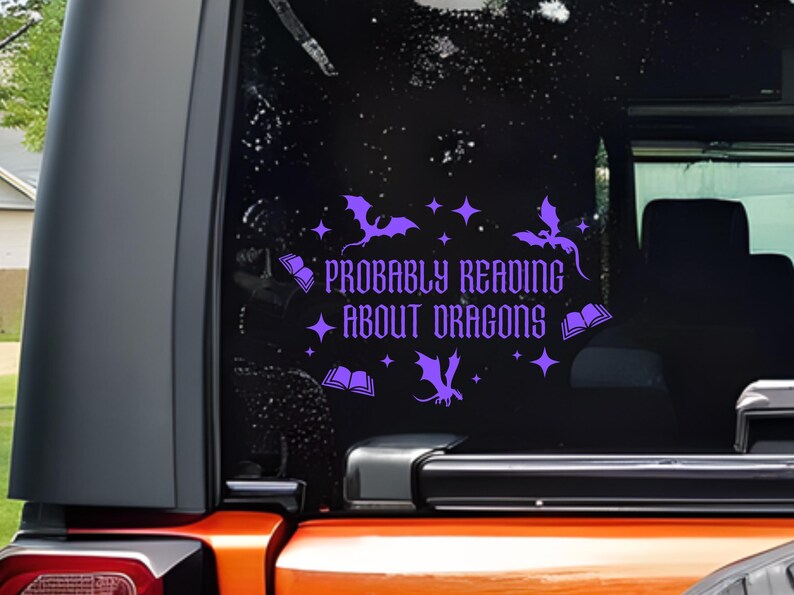 May include: Purple vinyl decal on a vehicle window, featuring the text "Probably Reading About Dragons" in a decorative font. The design includes dragon and book illustrations. The decal is applied to a black window.
