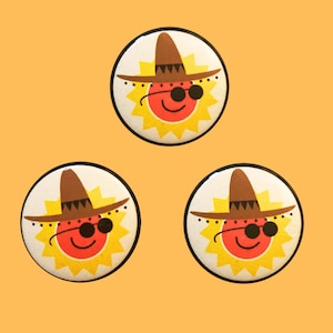 May include: Three round buttons with a cartoon sun wearing a brown sombrero and sunglasses. The sun has a red face and yellow rays.