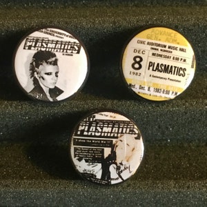 May include: Three black and white button badges featuring the band Plasmatics. The first badge shows a black and white photo of a woman with a mohawk. The second badge is a ticket for a Plasmatics concert on December 8, 1982. The third badge shows a black and white photo of a woman with a microphone.