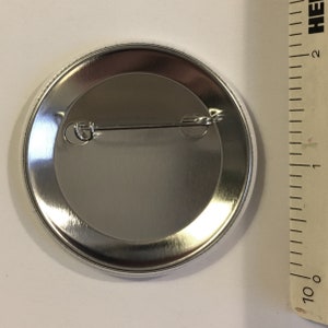K&ouml;nnte beinhalten: Die R&uuml;ckseite eines silbernen Knopfes mit einer daran befestigten Nadel. Der Knopf hat einen Durchmesser von 2,5 cm.