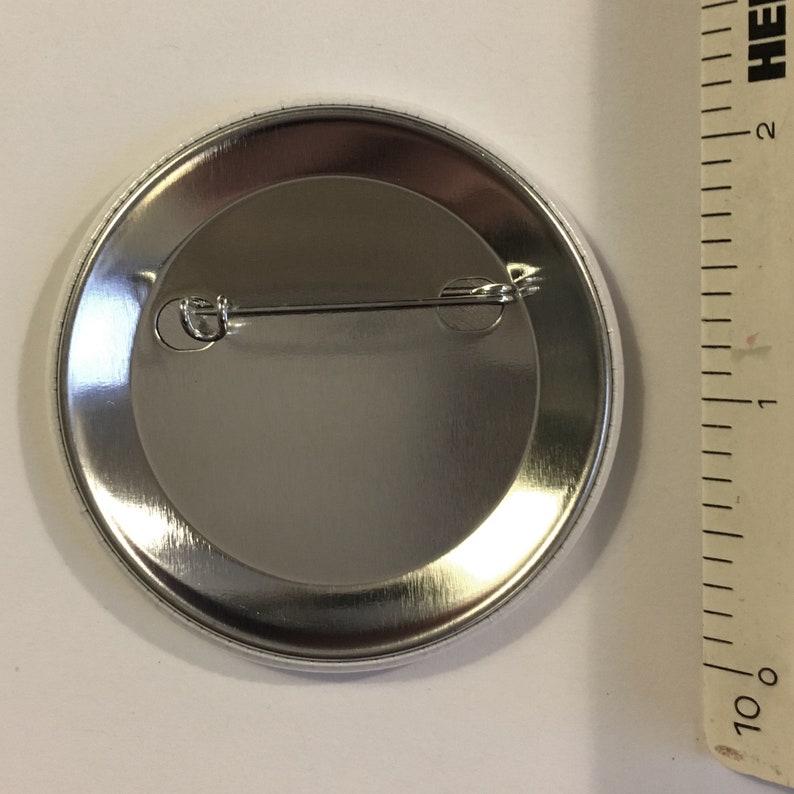 K&ouml;nnte beinhalten: Die R&uuml;ckseite eines silbernen Knopfes mit einer daran befestigten Nadel. Der Knopf hat einen Durchmesser von 2,5 cm.