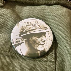 May include: A circular pinback button featuring a black and white graphic of a person wearing a military helmet with the text "MEAT IS MURDER" printed on the front. The button has a glossy finish and is displayed against a textured olive green fabric.