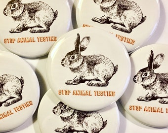 May include: Six round white buttons each featuring a black and white illustration of a rabbit. The buttons have the text "Stop Animal Testing" printed in orange on each.