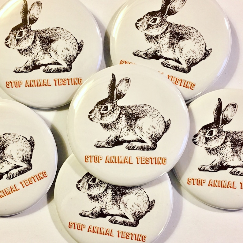 K&ouml;nnte beinhalten: Sechs runde wei&szlig;e Kn&ouml;pfe mit einer Schwarzwei&szlig;illustration eines Kaninchens auf jedem. Die Kn&ouml;pfe haben den Text "Stop Animal Testing" in Orange auf jedem.