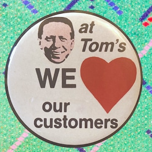 May include: A white button with a red heart and the text "WE ♥ our customers at Tom's" in black. A man's face is in the top left corner of the button.
