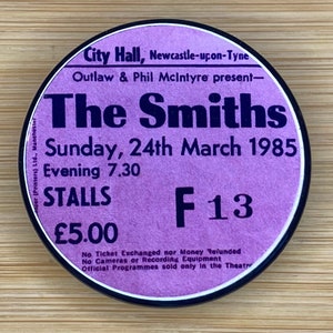 May include: A purple and black concert ticket for The Smiths, a British rock band, performing at City Hall in Newcastle-upon-Tyne on Sunday, March 24, 1985. The ticket is for the stalls section and costs £5.00. The seat number is F13.