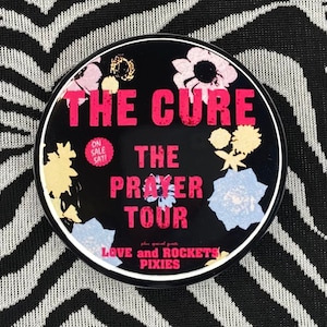 May include: A black and white circular button with a floral design. The text "THE CURE THE PRAYER TOUR" is in red. The text "LOVE and ROCKETS PIXIES" is in white.