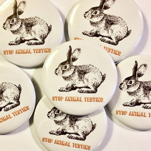 May include: Six round white buttons with a black and white illustration of a rabbit on each. The buttons have the text "Stop Animal Testing" printed in orange on them.