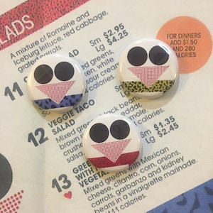 May include: Three round buttons with colorful geometric designs. Each button features a red, white, and black design with a different pattern. The buttons are on a white background with text that reads "VEGGIE TACO SALAD" and "GREEN WITH VEGETA".