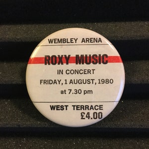 May include: A white button with black text that reads "Wembley Arena", "Roxy Music", "In Concert", "Friday, 1 August, 1980", "at 7.30 pm", "West Terrace", and "£4.00". A red stripe is above the text "Roxy Music".