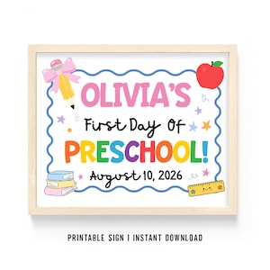 以下が含まれることがあります： 明るい木製のフレームに入った長方形の看板で、OLIVIA'S First Day Of PRESCHOOL! August 10, 2026という文字が書かれています。デザインには、ピンクのリボンが付いた黄色い鉛筆、赤いリンゴ、3冊の本の積み重ね、顔が描かれた黄色い定規が含まれています。