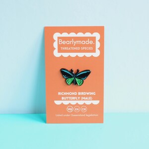 May include: A black and green enamel pin of a Richmond Birdwing Butterfly, a threatened species. The pin is on a card with the text "Bearlymade. Threatened Species" and "Richmond Birdwing Butterfly (Male)". The card also includes the conservation status of the butterfly: VU EN CR. The text "Listed under Queensland legislation" is at the bottom of the card.