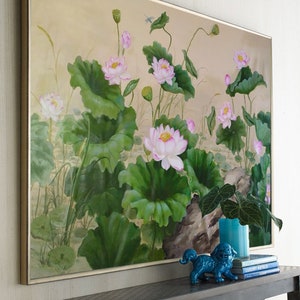 May include: A framed painting of pink water lilies with green leaves and stems on a beige background. The painting is hanging on a wall above a black console table with a blue ceramic figurine and books.