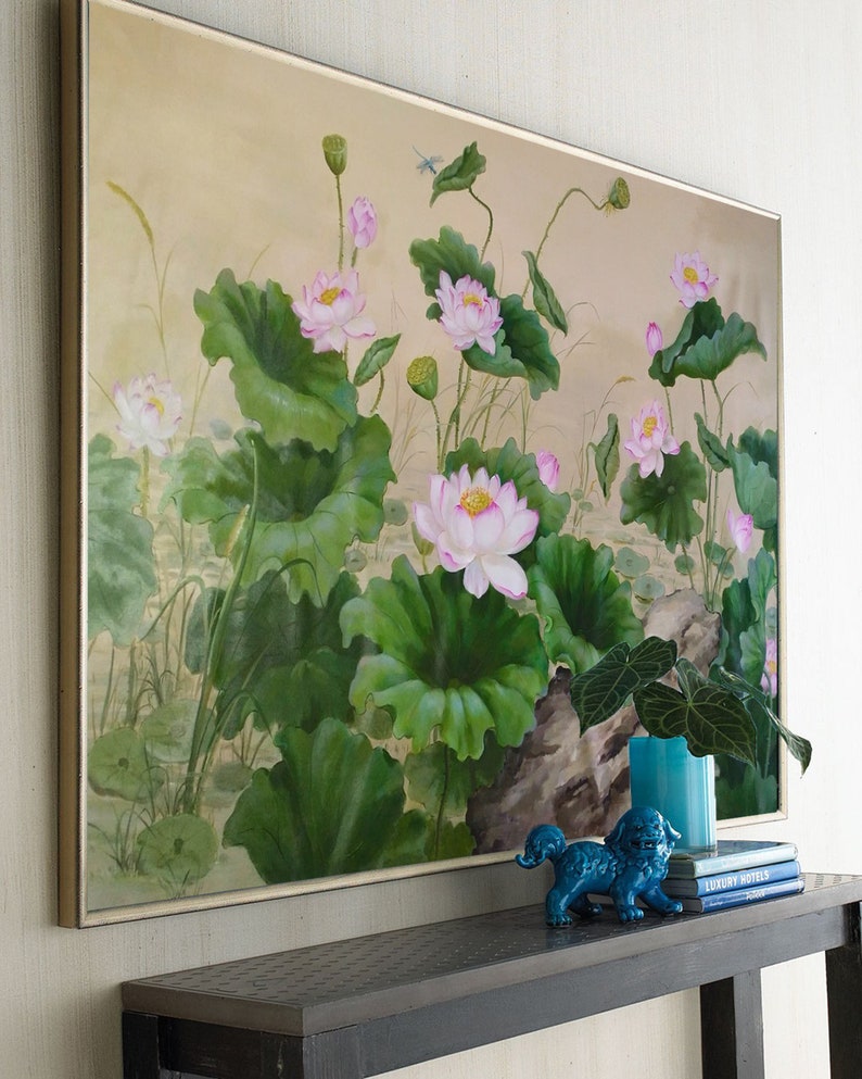 May include: A framed painting of pink water lilies with green leaves and stems on a beige background. The painting is hanging on a wall above a black console table with a blue ceramic figurine and books.
