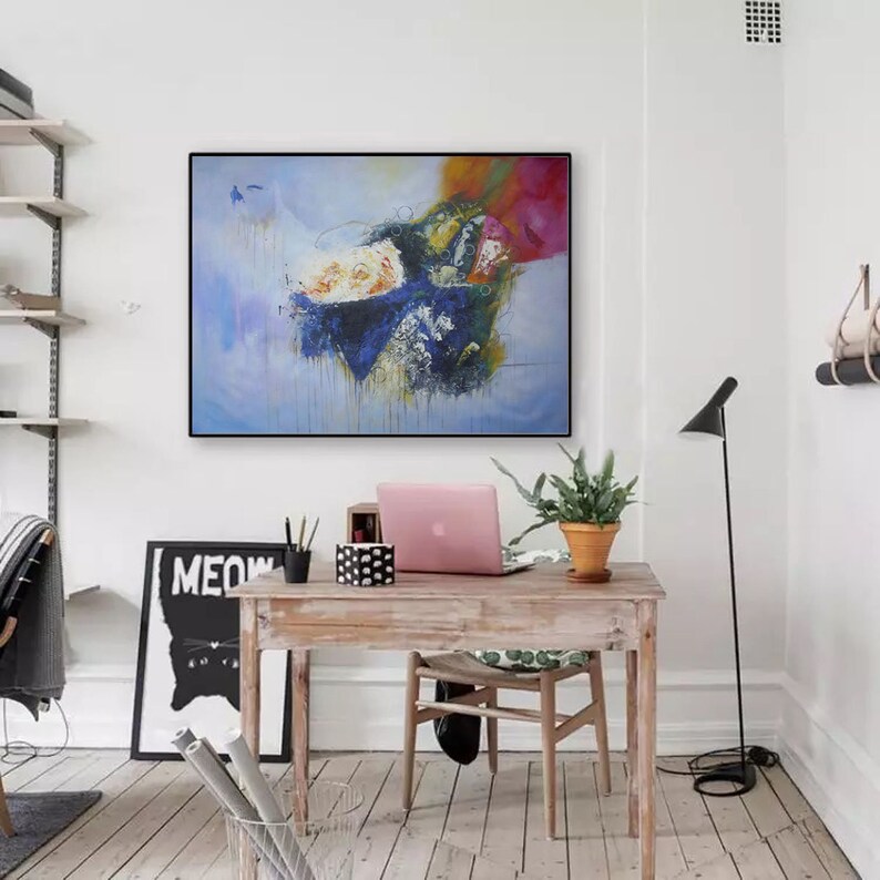 May include: Abstract painting with blue, white, and orange colors on a canvas. The painting is framed in black and hanging on a white wall above a wooden desk with a laptop, plant, and other office supplies.