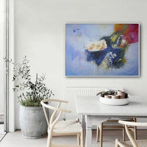 May include: Abstract painting with blue, yellow, white, and pink colors. The painting is framed in a white frame and is hanging on a white wall. The painting is in a living room setting with a white table and chairs.