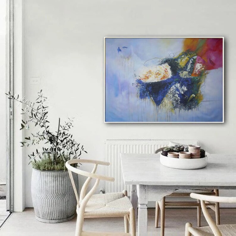 May include: Abstract painting with blue, yellow, white, and pink colors. The painting is framed in a white frame and is hanging on a white wall. The painting is in a living room setting with a white table and chairs.