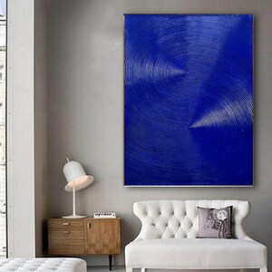 May include: Abstract painting with a blue background and concentric circles in white. The painting is hanging on a wall in a room with a white couch and a wooden side table.