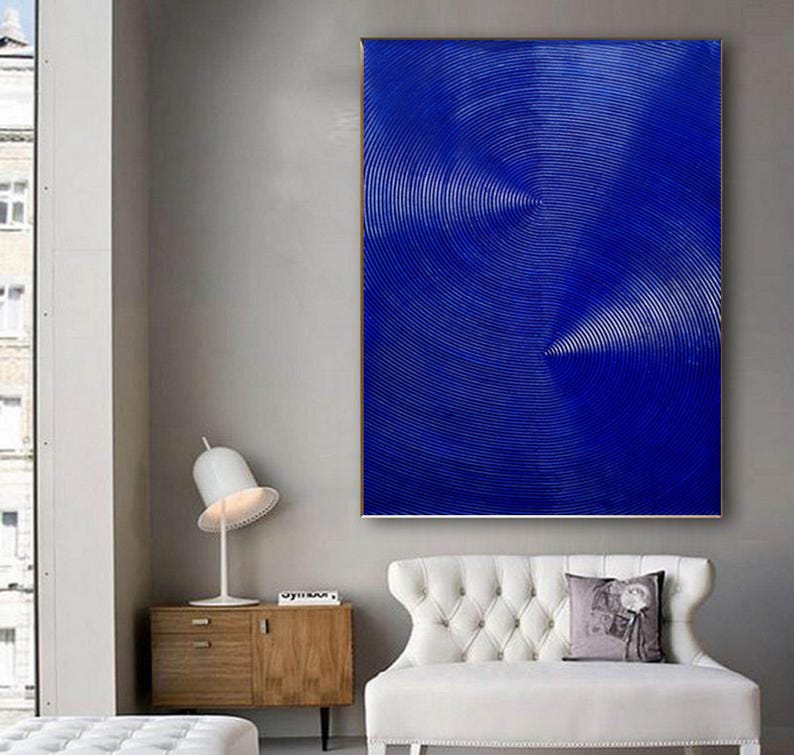May include: Abstract painting with a blue background and concentric circles in white. The painting is hanging on a wall in a room with a white couch and a wooden side table.