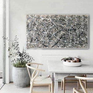 May include: Abstract black and white painting with a white background and black lines and splatters. The painting is framed and hanging on a white wall above a white table with chairs.