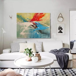 May include: Abstract painting with splashes of red, blue, green, and white paint on a yellow background. The painting is framed and hanging on a white wall above a white sofa.
