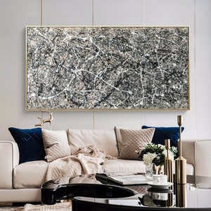 May include: Abstract black and white painting with a beige background, hanging on a white wall above a beige sofa with cushions and a coffee table.