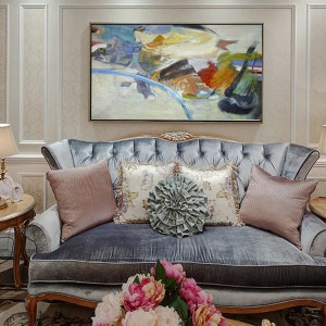 May include: Abstract painting with a mix of blue, green, yellow, orange, and red colors. The painting is framed and hanging on a wall above a gray velvet sofa with pillows.