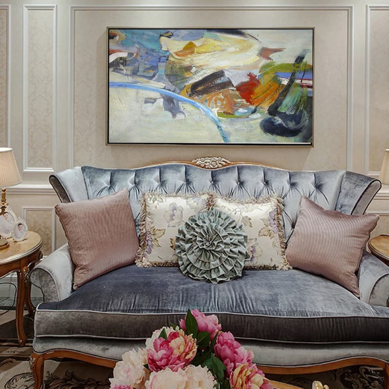 May include: Abstract painting with a mix of blue, green, yellow, orange, and red colors. The painting is framed and hanging on a wall above a gray velvet sofa with pillows.