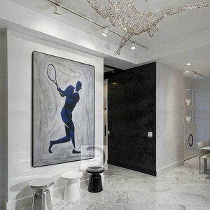 K&ouml;nnte beinhalten: Ein modernes Innendesign mit einem gro&szlig;en abstrakten Gem&auml;lde eines Tennisspielers in Blau und Wei&szlig; auf grauem Hintergrund. Das Gem&auml;lde h&auml;ngt an einer wei&szlig;en Wand mit drei silbernen und schwarzen Beistelltischen. Der Raum hat einen wei&szlig;en Marmorboden und eine schwarze Wand mit einer T&uuml;r, die zu einem anderen Raum f&uuml;hrt.