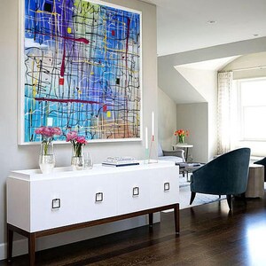 May include: A white cabinet with four drawers and brown legs sits in front of a large abstract painting with colorful lines and shapes on a blue background. The cabinet is in a room with hardwood floors and a view of a living room with a blue armchair.