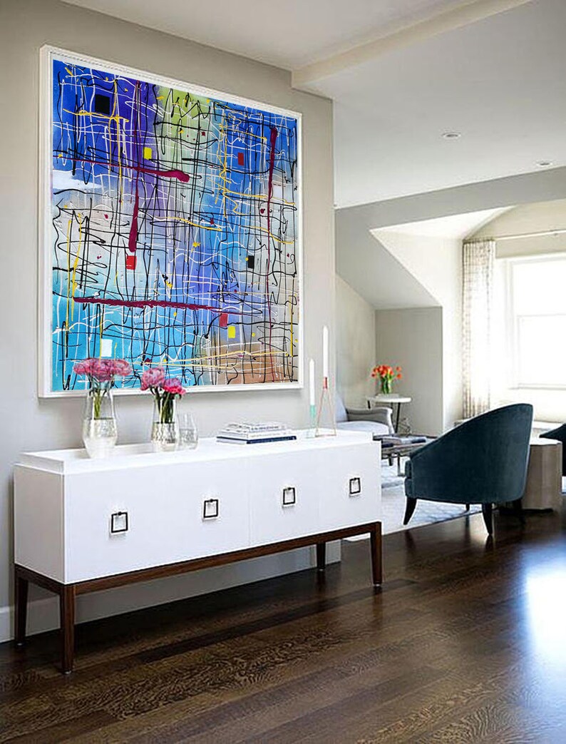 May include: A white cabinet with four drawers and brown legs sits in front of a large abstract painting with colorful lines and shapes on a blue background. The cabinet is in a room with hardwood floors and a view of a living room with a blue armchair.