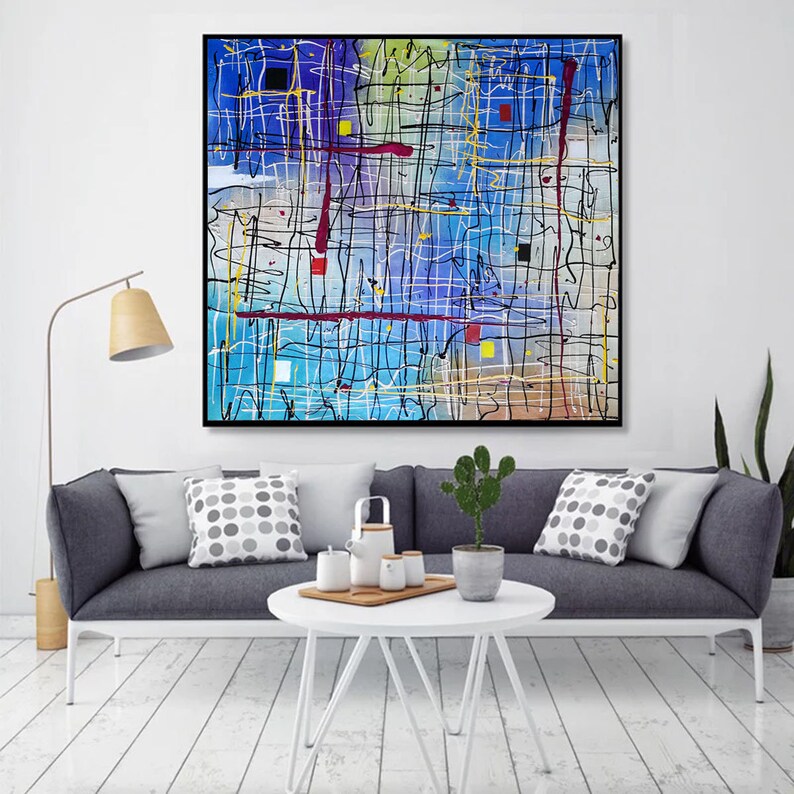 May include: Abstract painting with a blue background and black, white, red, and yellow lines. The painting is framed in black and hanging on a white wall above a gray couch with pillows.