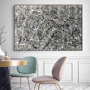 May include: Abstract painting with black, white and grey paint splatters and lines on a beige background. The painting is framed in black and hanging on a white wall.