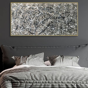 May include: Abstract black and white painting in a gold frame hanging above a grey striped bed with two pillows.