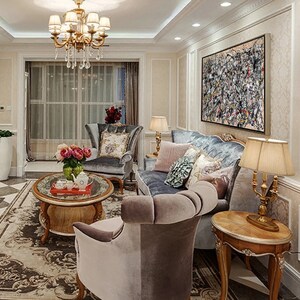 May include: A living room interior with a large abstract painting in black, white, and red hanging on the wall above a gray velvet sofa. The sofa is decorated with throw pillows and a throw blanket. There is a coffee table in the center of the room with a vase of flowers and a teapot on it. Two armchairs are positioned on either side of the coffee table. The room is decorated in a classic style with gold accents and a chandelier.