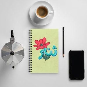 Könnte beinhalten: Ein spiralgebundenes Notizbuch mit hellgrünem Einband, das eine rote Blume und türkisfarbene arabische Schrift zeigt. Eine Tasse Espresso, ein Bleistift, ein Telefon und eine silberne Kaffeemaschine sind ebenfalls zu sehen.