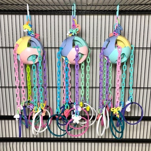 May include: Three colorful bird toys hanging from a cage. Each toy features a pastel-colored ball with a chain of links in various colors, including pink, purple, and green. The chains lead to a collection of rings and decorative charms.