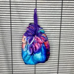Könnte beinhalten: Ein Kordelzugbeutel mit einem lebendigen Aquarellmuster in Blau-, Lila-, Rosa- und Gelbtönen. Der Beutel hat einen lila Kordelzug und eine passende Schlaufe oben. Der Beutel besteht aus einem weichen, möglicherweise samtartigen Material.