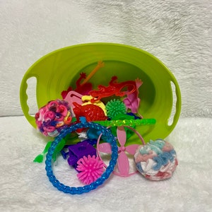 May include: A lime green plastic tub overflowing with colorful toys and accessories. The assortment includes pink and red plastic animal shapes, a blue coil bracelet, and two yarn balls. A variety of other small toys are also visible.