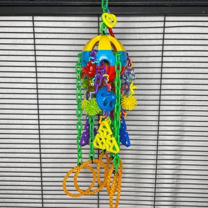 May include: A colorful bird toy with a yellow and blue dome, various plastic shapes, and hanging chains. The toy includes red hearts, green and purple shapes, and orange rings at the bottom. It is designed to hang inside a bird cage.