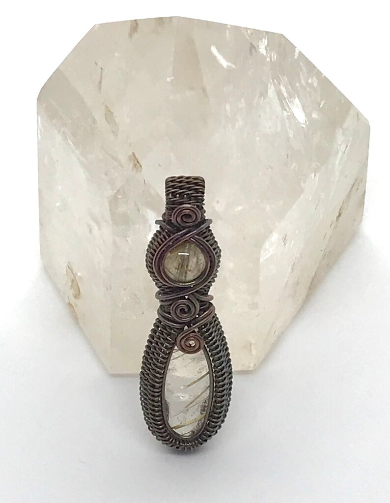 May include: A wire-wrapped pendant featuring two clear quartz stones with gold inclusions. The pendant is made with copper wire and has a rustic, antique finish.