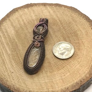 May include: A copper wire wrapped pendant featuring a clear quartz stone with a brown and gold iridescent sheen. The pendant is approximately the size of a US dime.