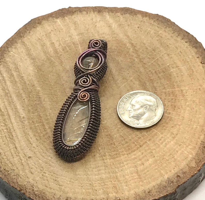 May include: A copper wire wrapped pendant featuring a clear quartz stone with a brown and gold iridescent sheen. The pendant is approximately the size of a US dime.