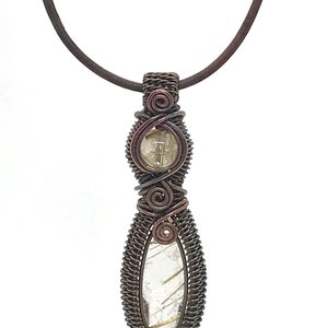 May include: A copper wire wrapped pendant necklace featuring a clear quartz crystal with gold inclusions. The pendant is suspended from a brown leather cord.