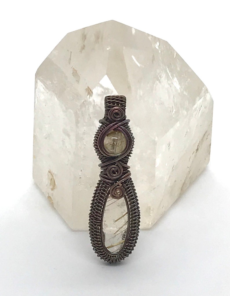 May include: A wire-wrapped pendant featuring a clear quartz crystal with a brown wire design. The pendant is displayed on a clear quartz crystal.