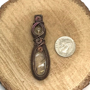May include: A copper wire wrapped pendant with a clear stone with brown and gold inclusions. The pendant is about the size of a US dime.