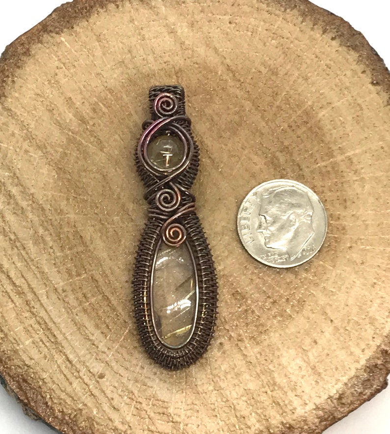 May include: A copper wire wrapped pendant with a clear stone with brown and gold inclusions. The pendant is about the size of a US dime.